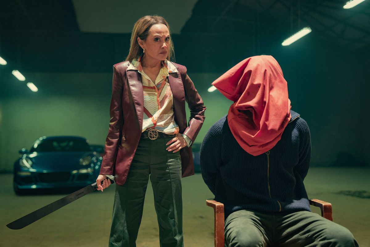 Martha Millan as Mercy in The Gentlemen holding a machete and standing next to a man tied to a chair with a bag on his head